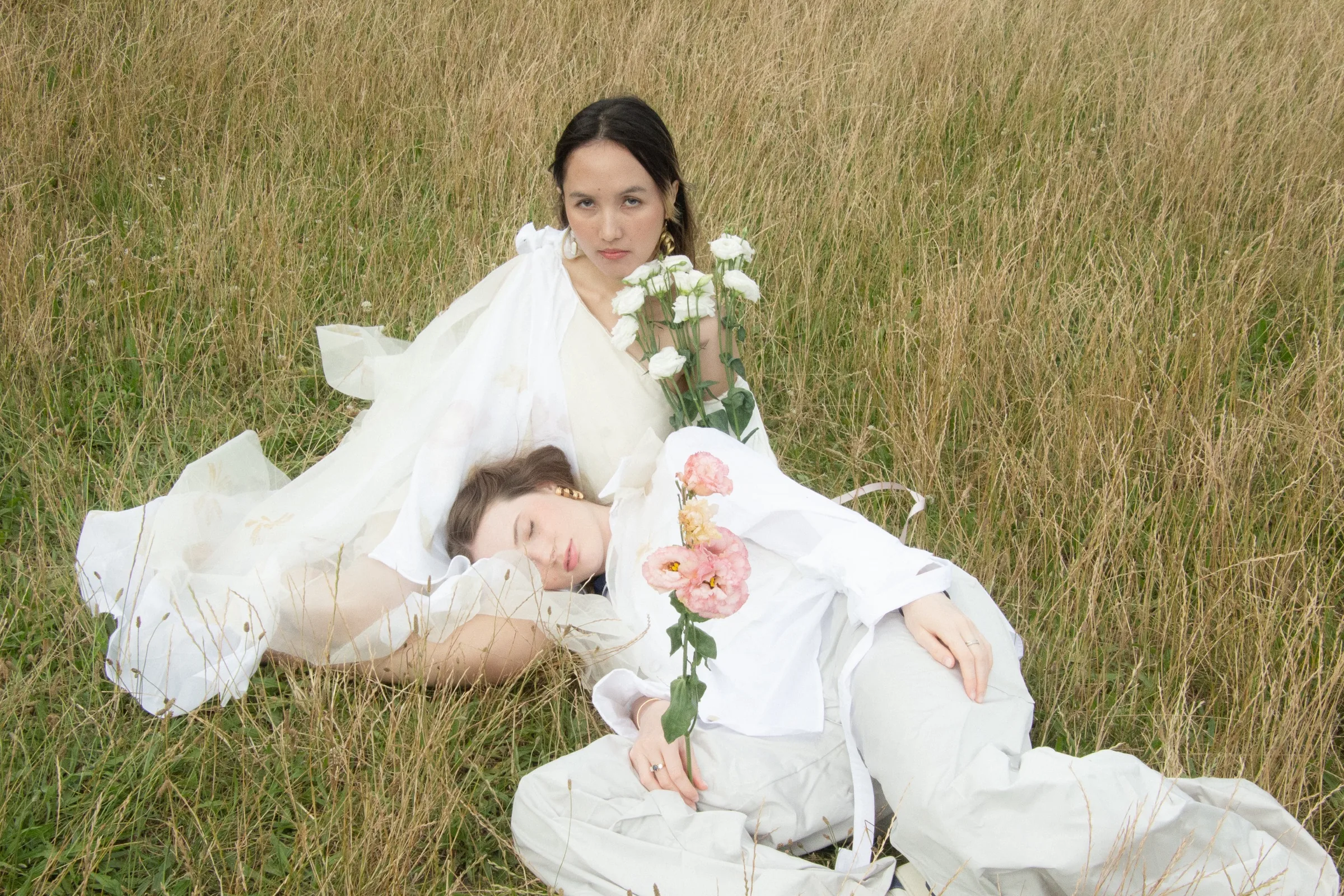 Two models lie in tall grass, one gazing upward, an armful of white blooms between them